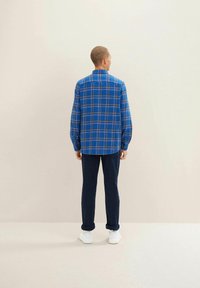 Niebieska koszula w kratę flanelowa z długimi rękawami, z zapięciem na guziki przy mankietach i prostym dołem, zestawiona z granatowymi spodniami i białymi trampkami.