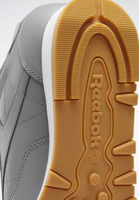 Pelēki sporta apavi ar gludu ādas virsotni, baltu vidusplāksni un teksturētu gumijas zoli. Iekļauta ieguļota logotipa un kontrastējoša šuvju dizains.