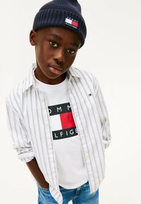 Striped white button-up shirt over a white t-shirt featuring a red and navy logo. Dark blue knit beanie on the head. Denim jeans.