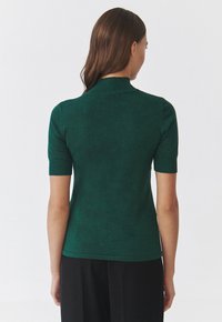 Zelený krátký rukáv top s úzkým střihem, texturovaným pleteným materiálem a límečkem polo, viděný zezadu, v kombinaci s černými kalhotami.
