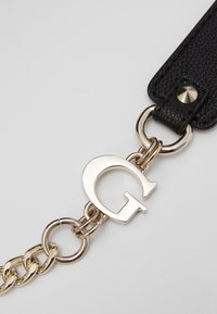 Cinturino in pelle nera texturizzata con catena in metallo tono oro e dettagli, con un grande ciondolo a forma di lettera "G" attaccato alla catena.