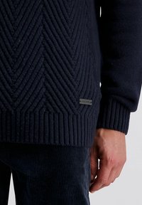Marineblauer Strickpullover mit einem strukturierten Zickzackmuster, gerippten Bündchen und einem kleinen Logoschild an der Seite.
