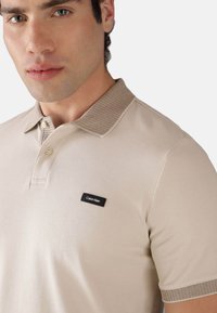 Polo beige con tessuto texture, colletto a costine in una tonalità più chiara e logo Calvin Klein nero sul petto.