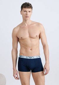 Άνδρας χωρίς πουκάμισο με κοντά, σγουρά μαλλιά, φορώντας μπλε σκούρα μποξεράκια με ανοιχτό γκρι ελαστική ζώνη που φέρει λογότυπο, στέκεται μπροστά σε απλό φόντο.