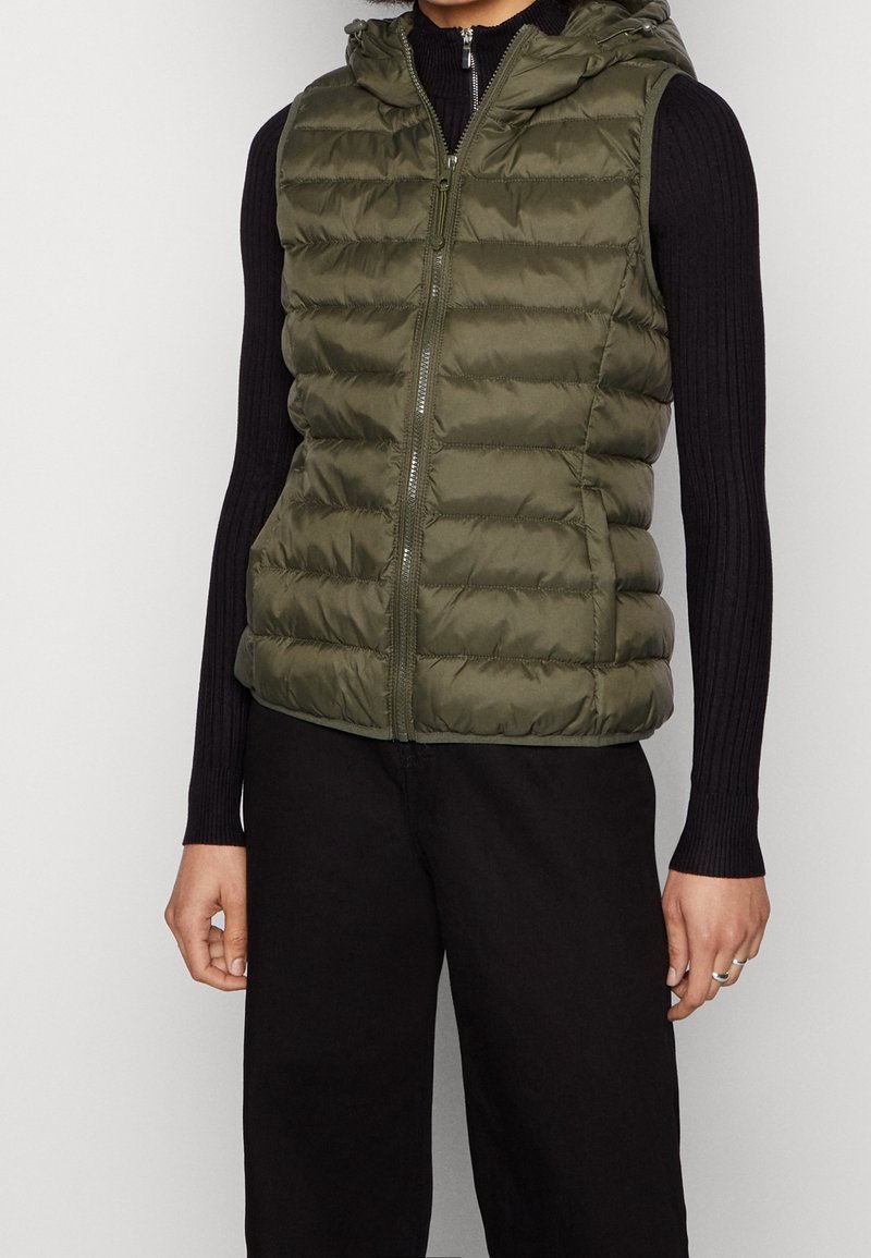 Gilet doudoune vert olive avec capuchon, doté d'une fermeture éclair à l'avant et d'une texture matelassée. Porté par-dessus un haut à manches longues noir à côtes.