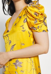 Femme portant une robe en satin jaune avec des manches bouffantes à motif floral et un décolleté en V profond, vue des épaules à la taille, portant des boucles d'oreilles créoles dorées.