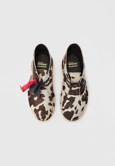Buty w brązowo-kremowy wzór w cętki krowy o zamszowej fakturze, z czarną metką i czerwonym akcentem. Wewnątrz: logo Clarks Originals Sandro.