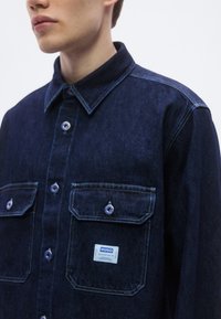 Σκούρο τζιν πουκάμισο με γιακά που κουμπώνει, δύο τσέπες στο στήθος και επώνυμη ετικ�έτα. Διαθέτει μεταλλικά κουμπιά και ραφές σε αντίθεση.