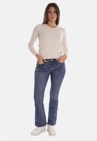 Maglione beige chiaro a maniche lunghe con scollatura a V abbinato a jeans in denim blu. I jeans hanno una vestibilità a gamba dritta e dettagli di cucitura discreti. Sneakers bianche.