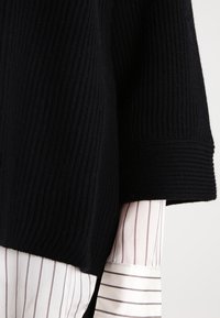 Schwarzer gerippter Strickpullover-Ärmel über dem weißen Hemdärmel mit dünnen vertikalen und horizontalen grauen Streifen.
