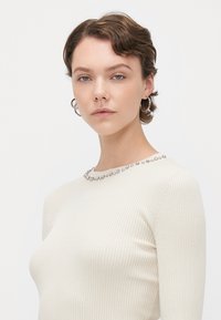 Ribbed, cream-colored long-sleeve top with a high neckline and decorative metallic beading around the collar. Smooth texture and fitted design.