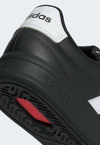 Gros plan sur le talon d'une basket Adidas noire et blanche avec une semelle noire texturée et un détail de bande de roulement rouge.