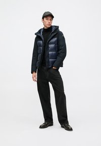 Doudoune avec capuche, bleu marine, présentant un design matelassé. Associée à un col roulé noir et un jean noir ample. Chaussures noires.