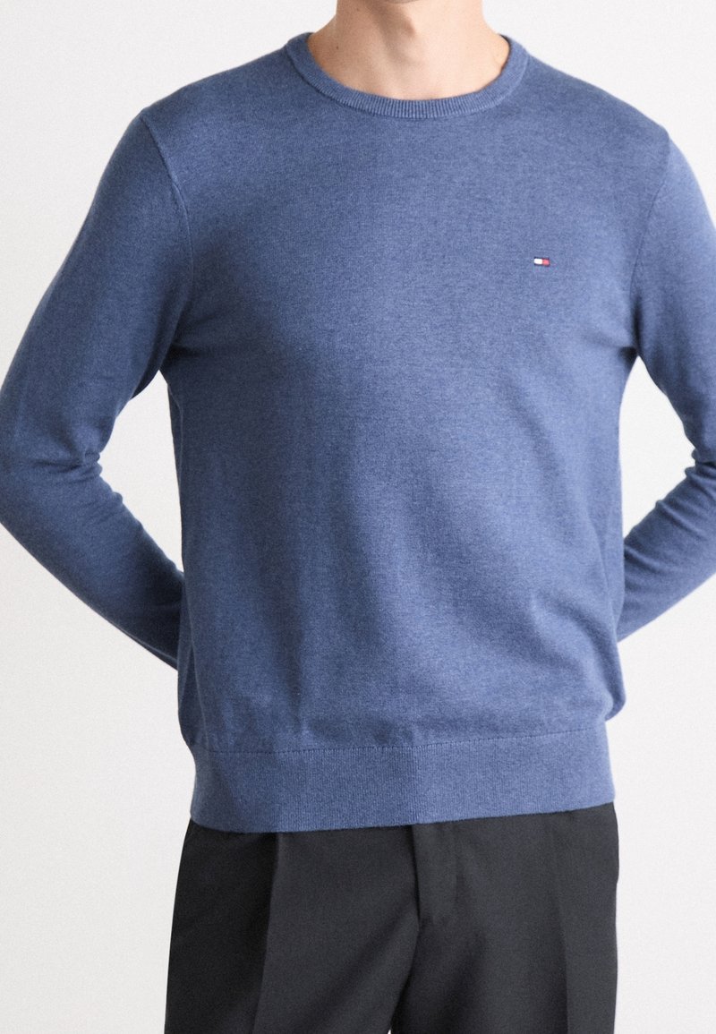 Homme portant un pull bleu à manches longues et col rond avec un petit logo de drapeau sur la poitrine et un pantalon noir.