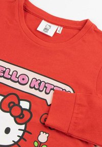 Red cotton long-sleeve shirt featuring a Hello Kitty graphic with a flower and text beneath. Ribbed neckline and cuff details visible.