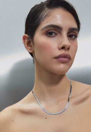 Žena s krátkymi tmavými vlasmi, ktorá nosí tenký strieborný náhrdelník a malý zlatý náušnicový manžet, pozerá sa mierne mimo objektív na jednoduchom pozadí.