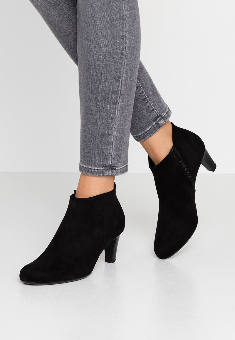 Jambes en jean skinny gris portant des bottines en daim noir avec fermetures éclair latérales et talons de hauteur moyenne sur un sol blanc.