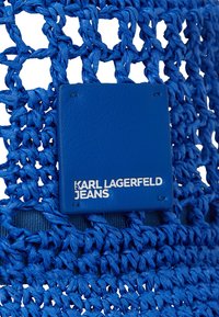 Modra pletena stolica iz teksturiranega materiala s kvadratno usnjeno oznako z napisom "KARL LAGERFELD JEANS" v belem tisku.