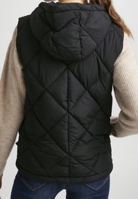 Czarna puchowa kamizelka z kapturem, z przeszyciami w diamentowy wzór i luźnym krojem, zestawiona z jasnobeżowym, teksturowanym swetrem pod spodem.