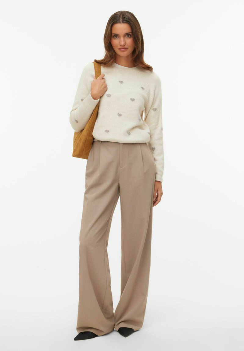 Pull crème à motifs de cœurs gris, associé à un pantalon beige à jambes larges et des chaussures noires pointues. Tenant un sac fourre-tout en daim marron.