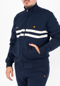 Giacca blu navy con zip e colletto alto, strisce bianche, logo sul petto. Materiale morbido, presenta tasche frontali e polsini a costine.
