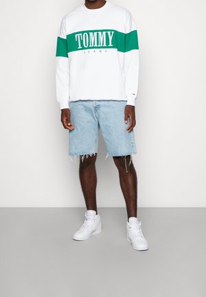 Uomo che indossa una felpa bianca con una banda verde e la scritta "TOMMY JEANS", pantaloncini di jeans azzurri sfrangiati e sneakers bianche alte.