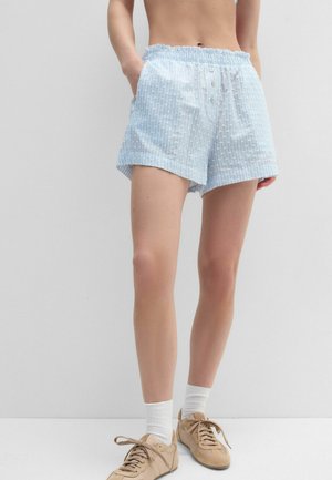 Persona que lleva puestos unos pantalones cortos texturizados azul claro con detalle de botón, calcetines blancos y zapatillas beige, de pie sobre un fondo liso.