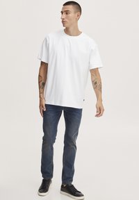 Man met kort haar, die een effen wit T-shirt, blauwe jeans, zwarte sneakers en zichtbare tatoeages op beide onderarmen draagt, staande en naar rechts kijkend.