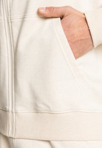 Light beige zip-up hoodie made from soft fabric. Features a front pocket and ribbed hem with stitching details. Hand resting in pocket.