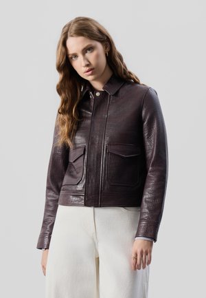 Femme aux longs cheveux ondulés portant une veste en cuir texturée marron foncé et un pantalon écru, debout devant un fond clair uni.