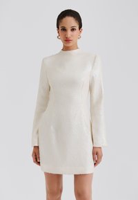 Krátke priliehavé šaty v krémovej farbe s dlhými rukávmi, vysokým golierom a textúrovaným flitrovaným povrchom so zakončením v tvare vlniek.