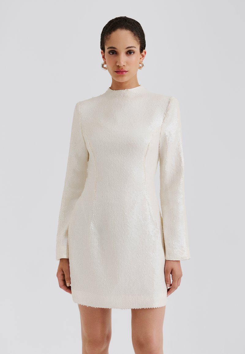 Krátke priliehavé šaty v krémovej farbe s dlhými rukávmi, vysokým golierom a textúrovaným flitrovaným povrchom so zakončením v tvare vlniek.