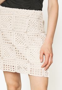 Mini spódnica na szydełku w jasnobeżowym kolorze, z kwadratowymi wzorami, teksturowanym splotem oraz elastycznym paskiem w talii dla dopasowania. Detale obejmują ażurowy wzór.