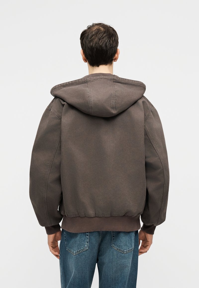 Persona che indossa una felpa con cappuccio marrone oversize e jeans blu, vista da dietro su uno sfondo neutro.