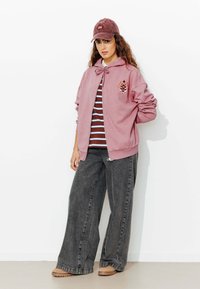 Sweat à capuche zippé rose avec un détail en nœud et un design brodé sur une chemise rayée, associé à un pantalon large gris et des chaussures beiges.