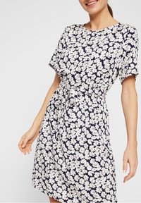 Woman wearing a short-sleeve navy dress with white floral print and a waist tie, smiling with a relaxed pose.