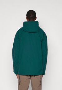 Zelena vodootporna jakna s kapuljačom, slobodnog kroja i suptilnom teksturom. Ima ojačane šavove i dizajn sa produženim repom za dodatnu zaštitu.