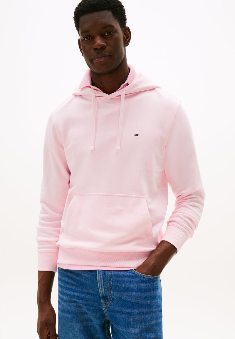 Felpa con cappuccio rosa chiaro realizzata in un tessuto morbido, caratterizzata da una tasca frontale, polsini a costine e un piccolo logo sul petto. Abbinata a jeans blu.