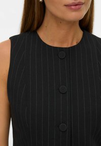 Crna haljina na crte bez rukava, s okruglim dugmadi prekrivenim tkaninom, glatkom teksturom i uskim siluetom s okruglim izrezom.