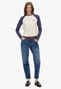 Kobieta ubrana w kremową i granatową koszulkę baseballową z napisem "Superdry", niebieskie dżinsy z brązowym paskiem, białe skarpetki i ciemnobrązowe mokasyny, stoi z rękami w kieszeniach.