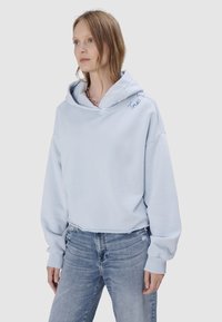 Kobieta w jasno niebieskiej krótkiej bluzie z kapturem na głowie i haftem "True" przy ramieniu, w połączeniu z wyblakłymi niebieskimi jeansami na białym tle.