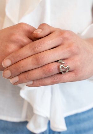 Handen in elkaar gevouwen, met een zilveren open hartvormige ring aan de ringvinger, witte kleding en een achtergrond van blauwe spijkerbroek.