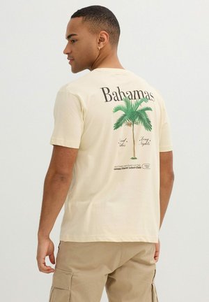 Joven de perfil con una camiseta beige que tiene una palmera verde y el texto "Bahamas" en la parte trasera, combinado con pantalones cortos cargo color caqui.