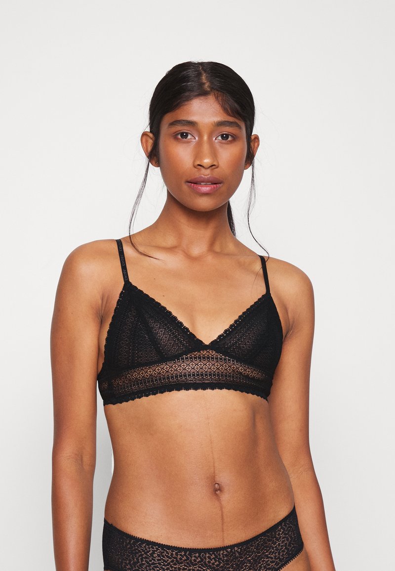 Giovane donna con capelli scuri che indossa un reggiseno di pizzo nero e biancheria intima coordinata, in piedi davanti a uno sfondo bianco semplice.