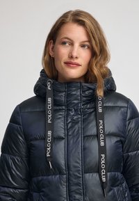 Marinblå puffjacka med horisontellt kviltat mönster, huva och svarta dragsnören med "POLO CLUB" branding. Glänsande, slät textur.