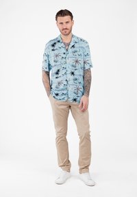 Uomo con tatuaggi in piedi, che indossa una camicia a maniche corte con stampa di palme blu, pantaloni beige e scarpe da ginnastica bianche su uno sfondo bianco.