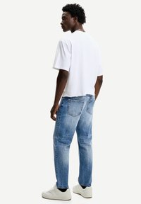 Witte katoenen t-shirt, ontspannen pasvorm, met korte mouwen. Vervaagde blauwe jeans met rechte snit, zichtbare achterzakken en versleten textuur. Witte sneakers.