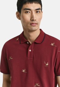 Junger Mann trägt ein burgunderfarbenes Poloshirt mit kleinen gestickten fliegenden und stehenden Fasanen, dunkle Akzente an Kragen und Ärmeln.