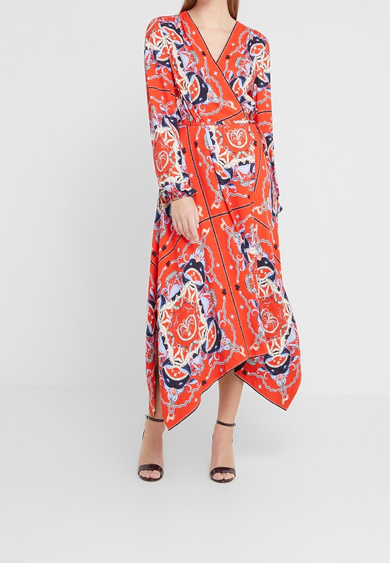 Femme portant une robe portefeuille orange à manches longues avec des motifs ornés bleus et blancs, un ourlet asymétrique et des talons noirs à bride cheville.
