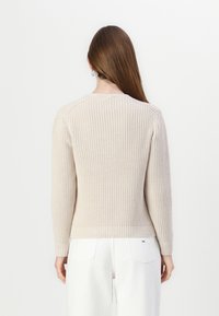 Pull en tricot beige à manches longues, avec un motif texturé et un ourlet côtelé, porté avec un pantalon blanc, vu de dos.